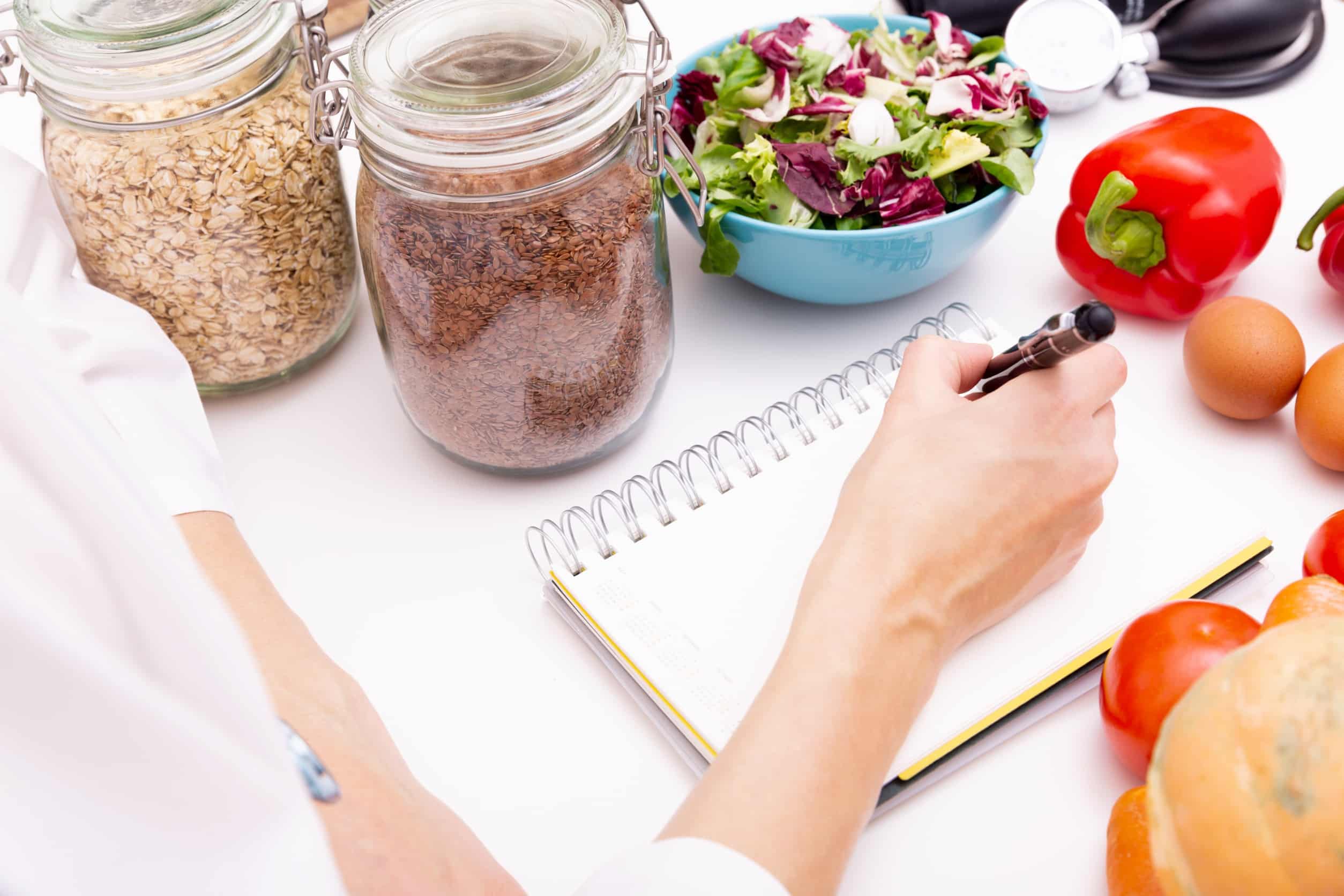 Les mains d'un nutritionniste écrivant dans un calepin posé sur un bureau avec des bocaux remplis de céréales, un bol de salade, des œufs et des légumes.