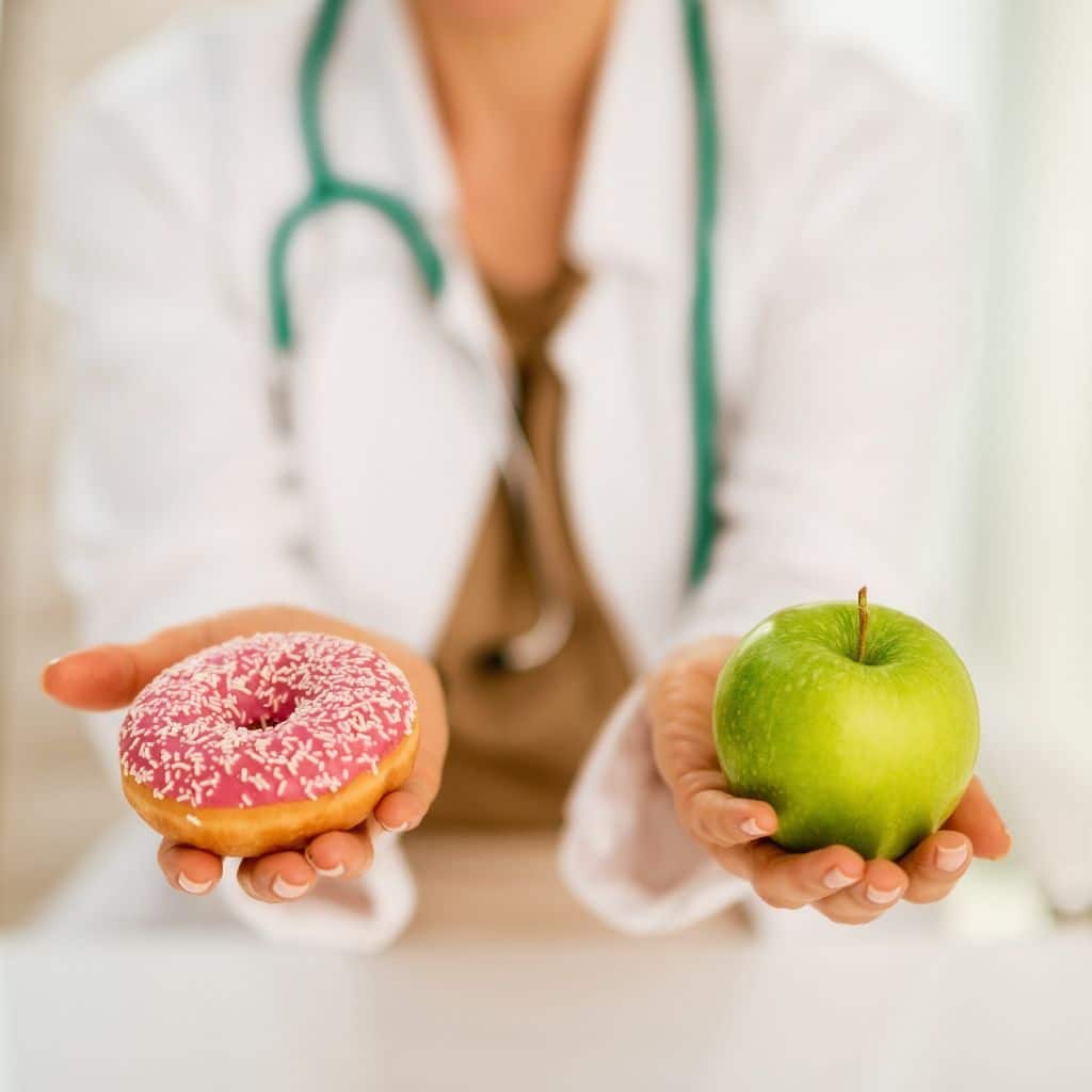 diététicienne en blouse tenant un donut de sa main droite et une pomme verte de sa main gauche. Vue de face sur les mains