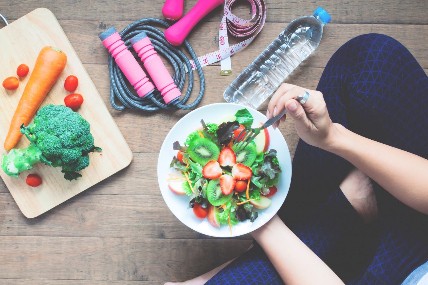 Une personne tenant une assiette contenant des aliments frais et sains, assise devant une corde à sauter, une bouteille d'eau, un mètre ruban et une planche avec des légumes.